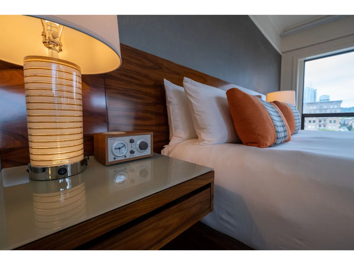 A plush guestroom scene featuring a cozy bed with decorative pillows, a lamp and nightstand, and a window letting in natural light. The mood is tranquil, suggesting comfort, relaxation, and the restorative ambiance promised to wellness-focused guests.