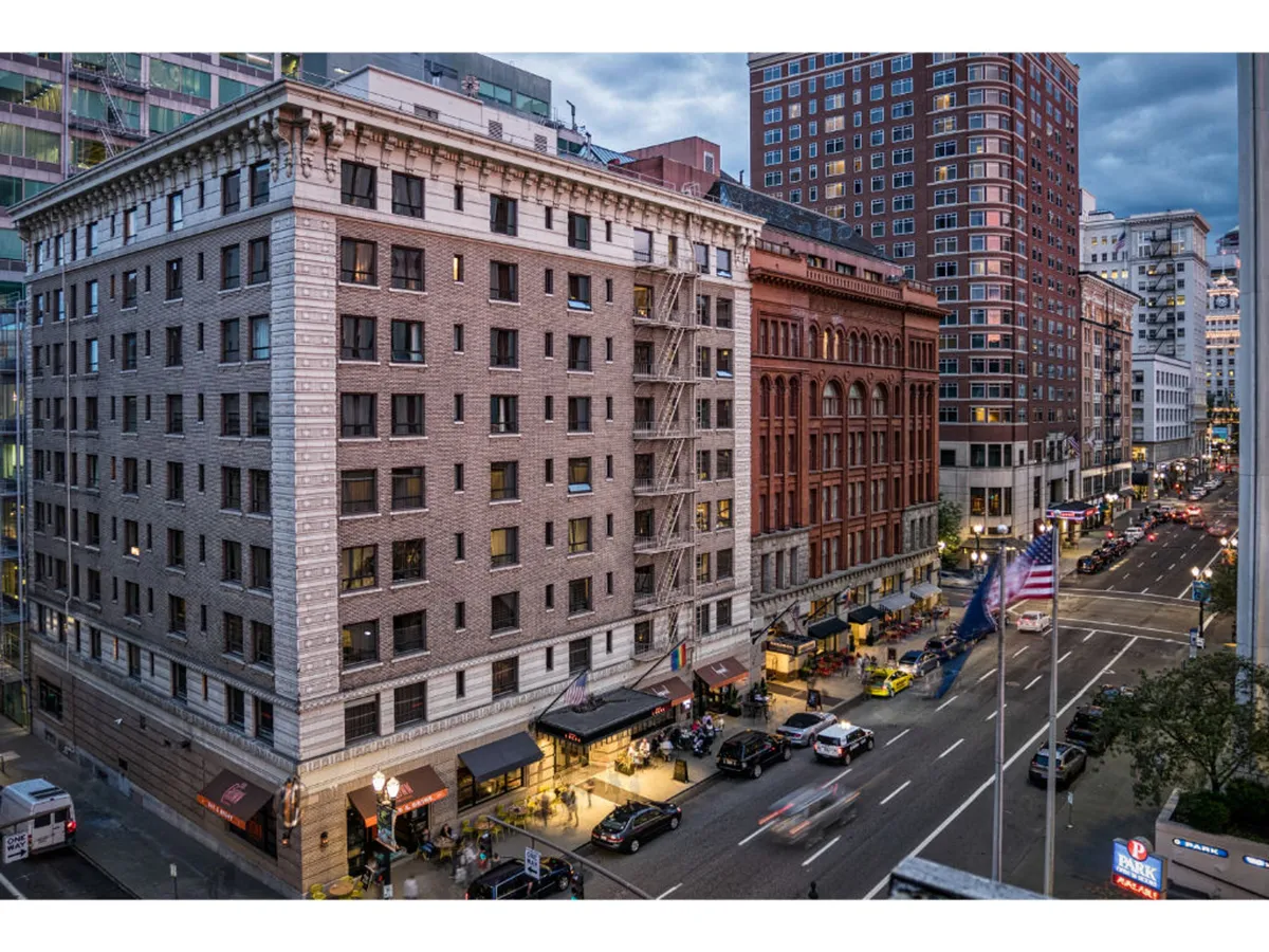 A bustling downtown Portland street scene with historic buildings, lively street activity, and restaurant awnings. People are out dining, shopping, and enjoying the vibrant energy of the city. The image evokes the dynamic cultural and social opportunities that await just steps from the hotel.