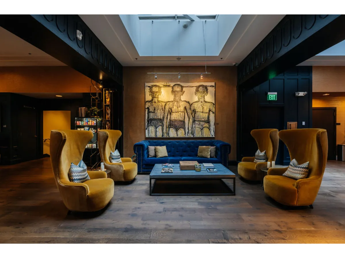 A stylish hotel lounge interior with a plush blue velvet couch, bright yellow armchairs, and striking artwork. The space is designed for relaxation, casual meetings, or unwinding after a busy day, with an inviting and contemporary ambiance.