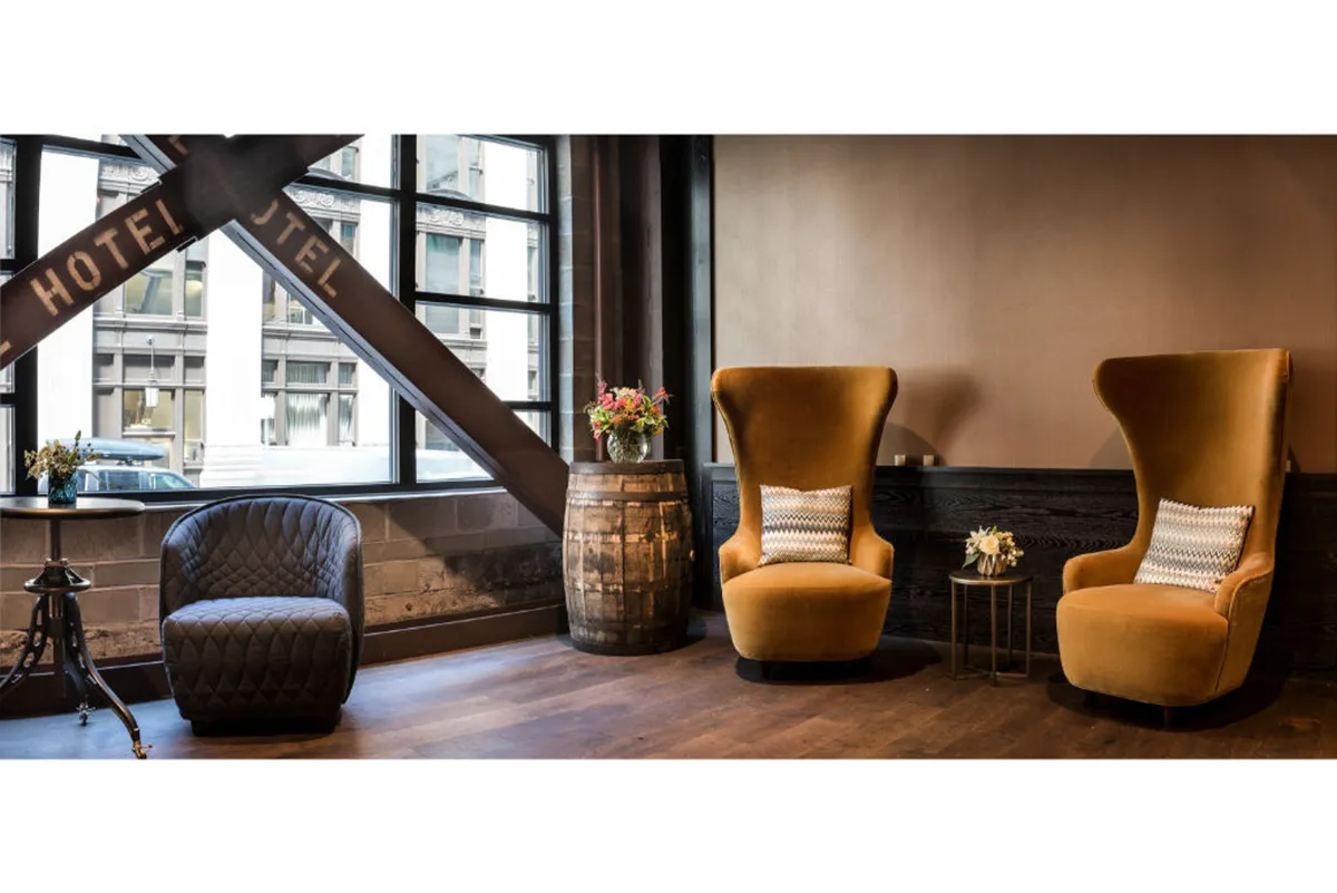 A cozy seating area featuring two large golden velvet chairs, a small black quilted chair, and a rustic wooden barrel. A vibrant flower arrangement sits atop a small table, and the space is warmly lit, giving it a welcoming, social feel.