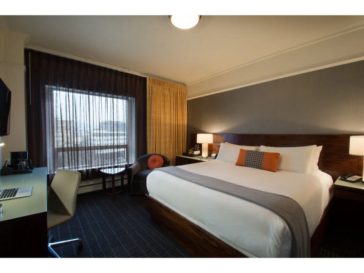A spacious Premium King Room featuring a large bed with crisp linens, a window with elegant curtains letting in natural light, a desk with a laptop for work or leisure, and a comfortable seating area. The design fuses historic architectural elements with contemporary furnishings, creating an inviting and stylish retreat.