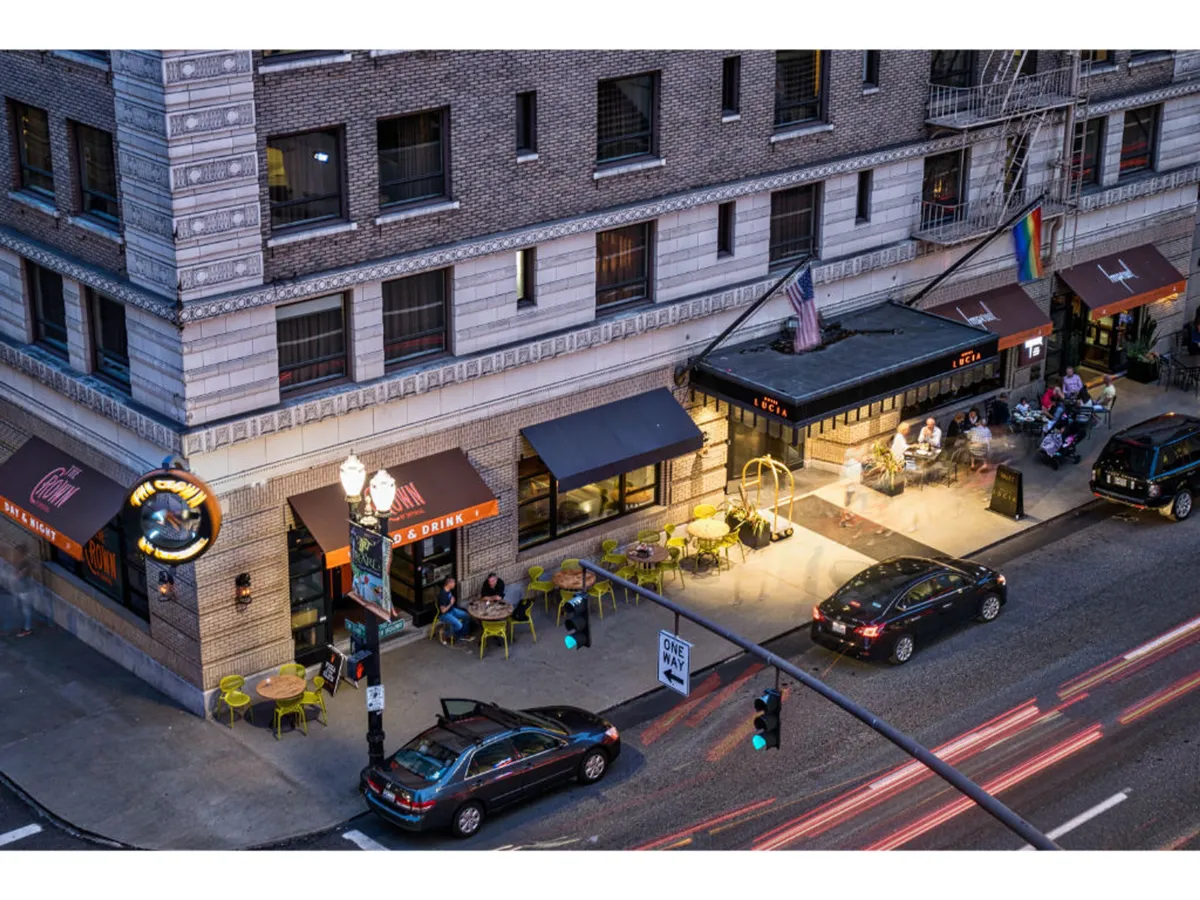 A lively exterior scene featuring the signage of a restaurant, inviting outdoor seating, busy city street traffic, and surrounding urban architecture. The image captures the energy and accessibility of downtown Portland, conveying a sense of being at the heart of the city’s vibrant activity.