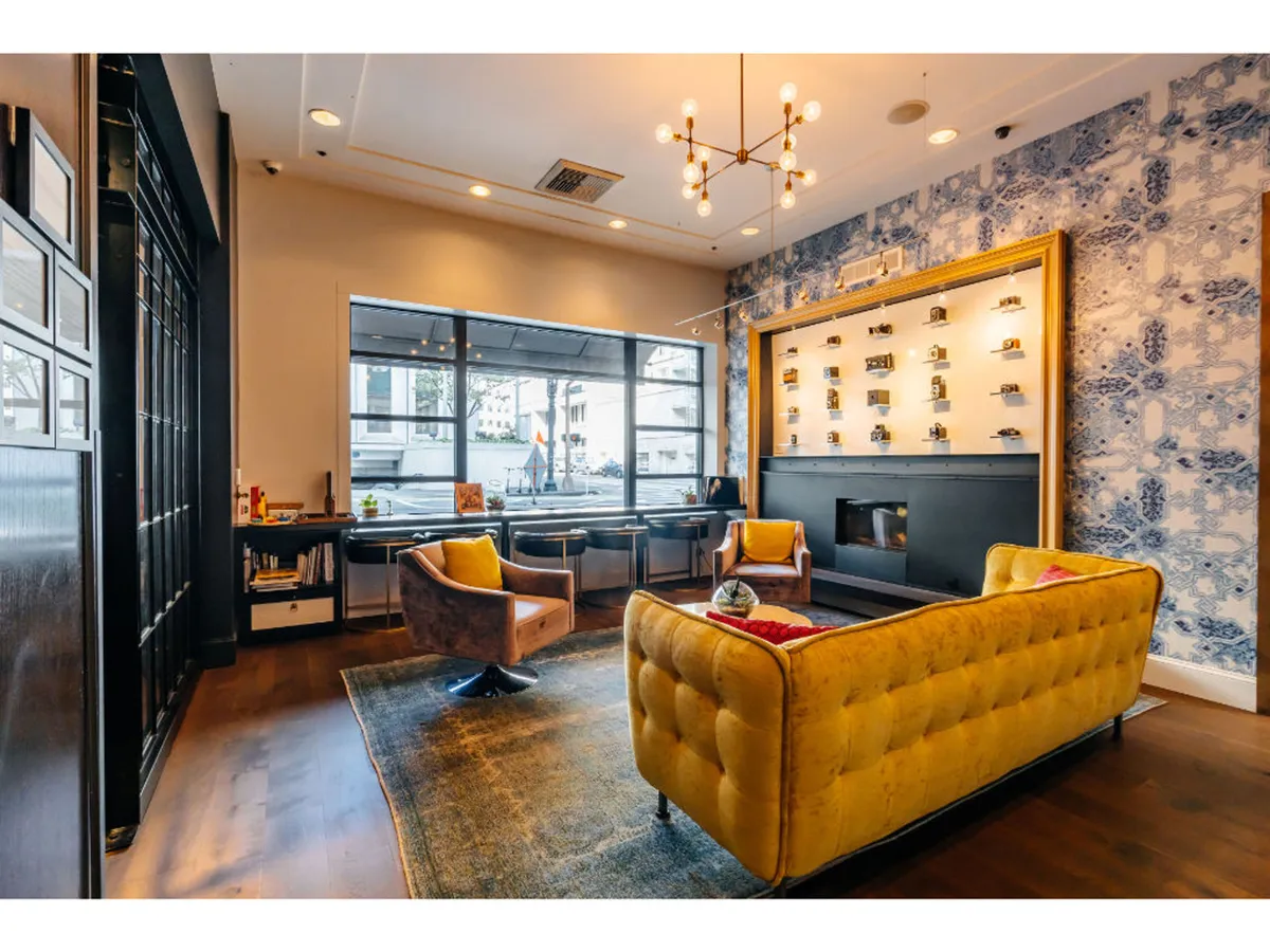 A welcoming lounge area with a vibrant yellow tufted sofa, two armchairs, and a decorative wall with open shelving. The space is ideal for casual meetings, reading, or engaging in lively conversation, evoking a sense of playful social connection.