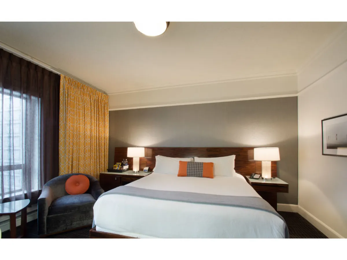 The image showcases a serene hotel room interior bathed in soft natural light, featuring a plush bed adorned with decorative pillows, elegant side lamps casting a warm glow, and a cozy armchair positioned by the window—inviting moments of relaxation or quiet reflection. The overall atmosphere is calm and welcoming, with thoughtful touches that blend comfort and style.