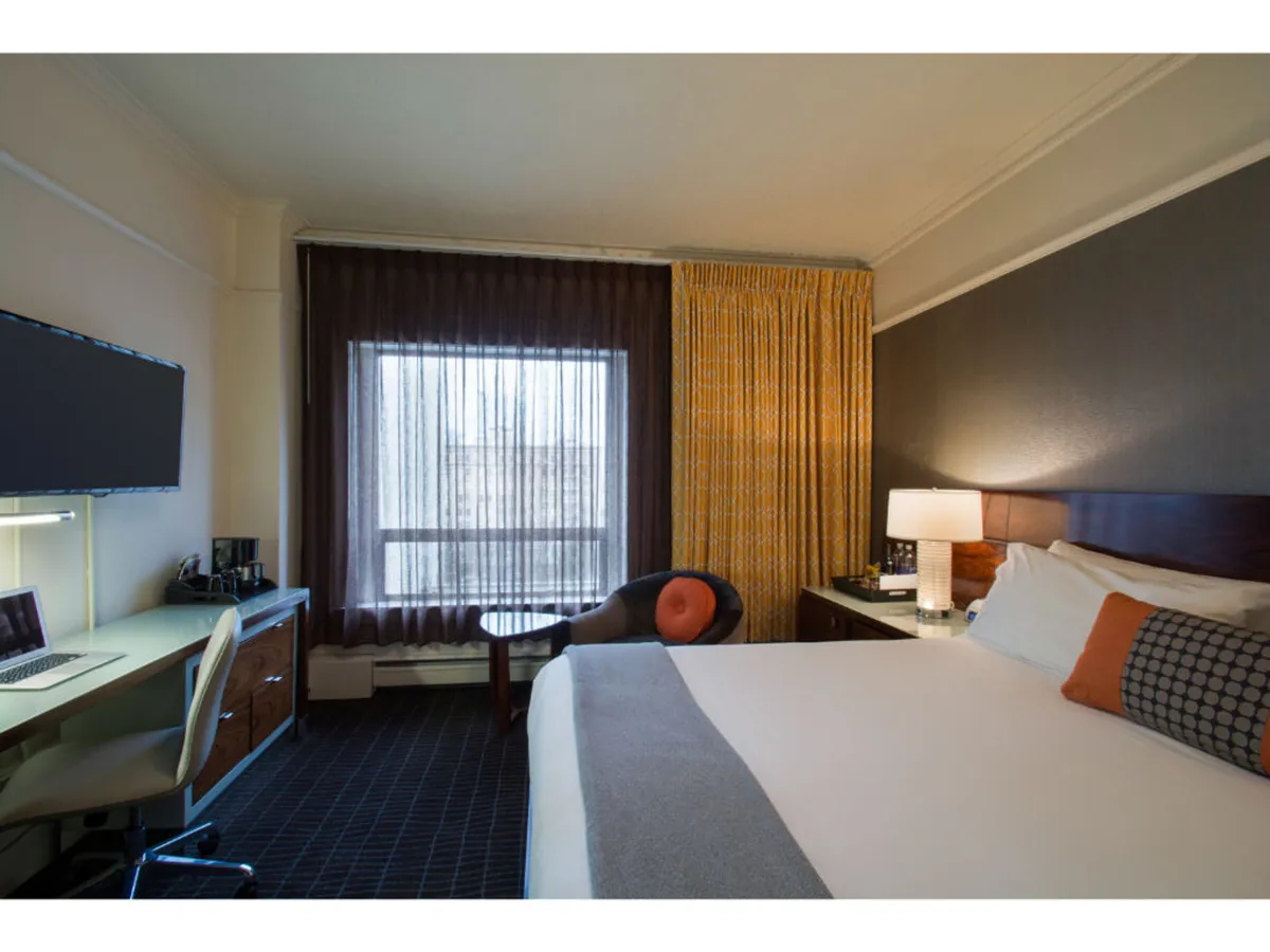 A contemporary guest room with a large bed, a desk equipped with a computer, an armchair, and a window with sheer curtains letting in natural light. The space feels tranquil and well-appointed, perfect for relaxing or catching up on work without worry.